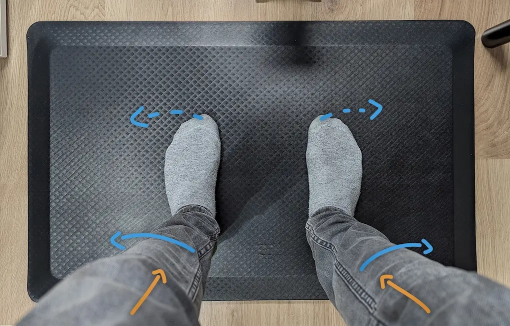 Widok z góry na stopy na macie antyzmęczeniowej z nałożonymi niebieskimi strzałkami instruującymi poprawną rotację bioder i stóp